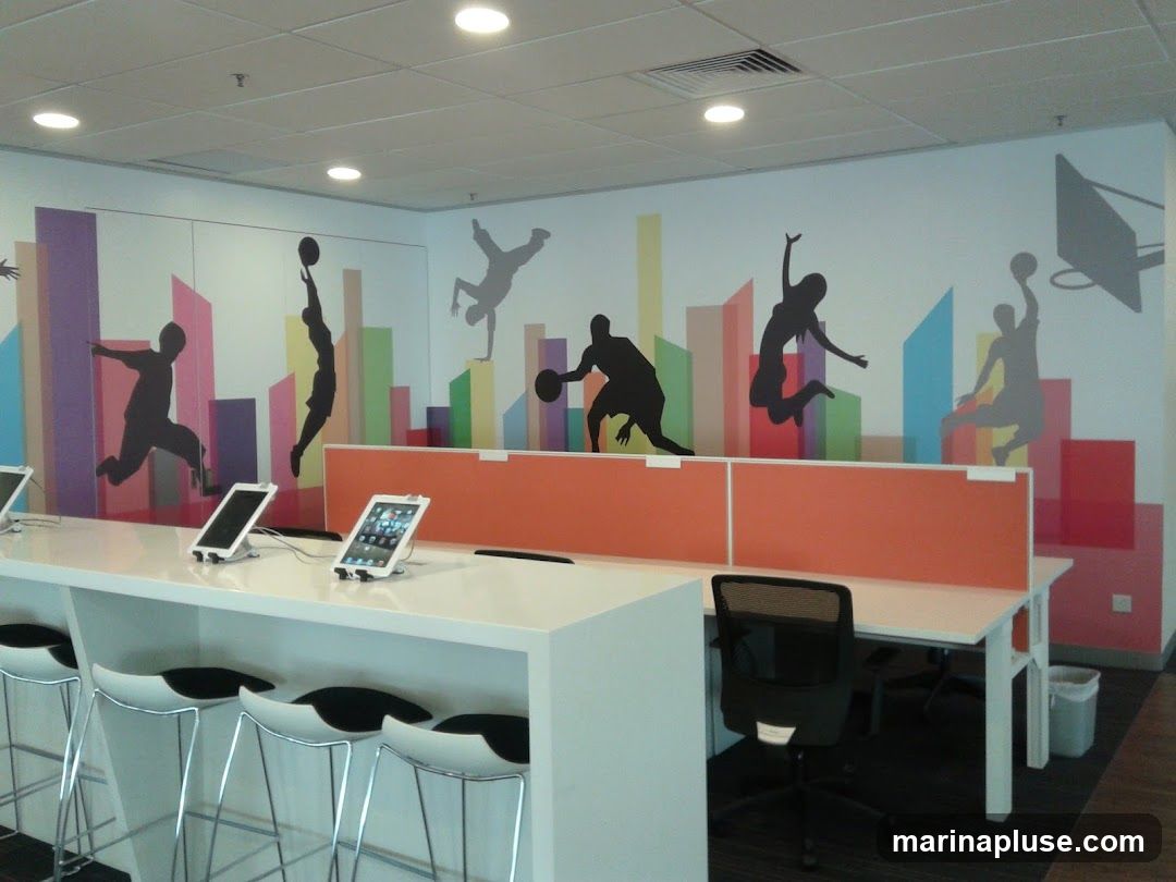 An interior view of a modern office space featuring a vibrant mural on the wall depicting silhouettes of people playing basketball and breakdancing against a backdrop of colorful abstract shapes resembling buildings. A long white counter with several stools and tablets is in the foreground. Orange partitions divide desk areas, and a black office chair is visible.