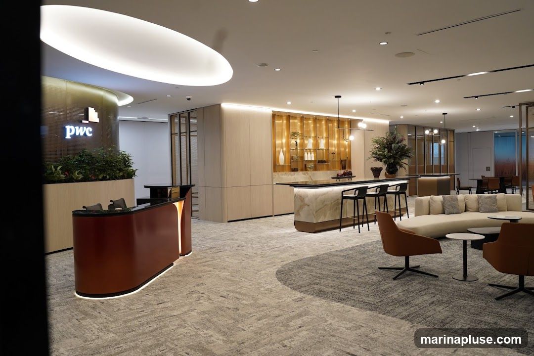A modern and spacious reception area with a prominent PwC logo. The space features a sleek reception desk, a marble-topped bar with stools, comfortable lounge seating, and decorative shelving. The overall ambiance is sophisticated and professional, with warm lighting and contemporary design elements.