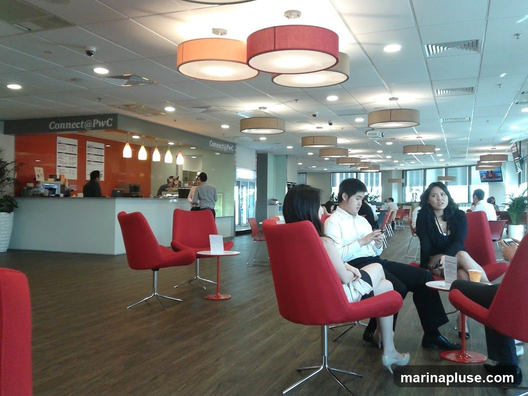 An interior view of a modern office break area, featuring a counter with staff serving customers. Several people are seated in red chairs, some engaged with their phones. The space is well-lit with overhead lighting and decorated with contemporary pendant lamps. The signage "Connect@PwC" is visible on the counter area.