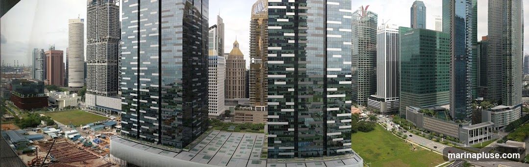 A panoramic view of a modern cityscape dominated by numerous tall skyscrapers of varying architectural styles. Some buildings are sleek and glass-covered, while others have more traditional facades. In the foreground and mid-ground, there are areas of green space, roads with traffic, and ongoing construction sites with cranes and building materials. The background shows a port or industrial area with cranes and ships, under a cloudy sky.