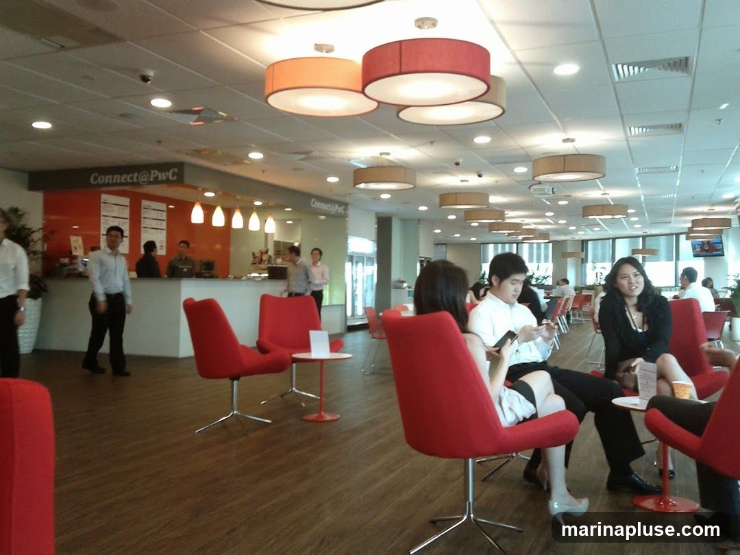 A modern office break area with a reception desk, seating, and people interacting. The reception desk has a sign that says "Connect@PwC". There are several red chairs arranged in the space, and people are seated and standing. The lighting is bright, with pendant lights and ceiling lights.