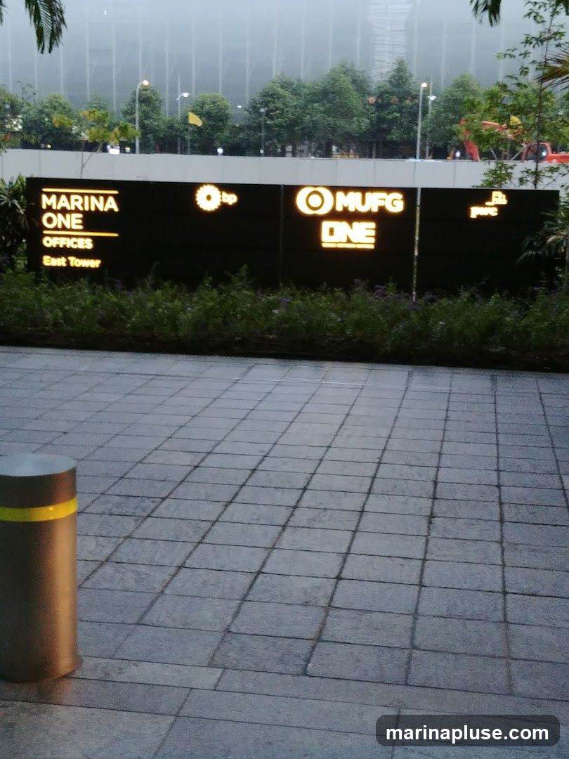 A dark sign with illuminated text and logos is mounted on a wall, with lush green bushes in front of it. The sign reads "MARINA ONE OFFICES East Tower" on the left, followed by the BP logo, then "MUFG ONE" with a circular logo, and finally "pwc" with a logo. In the background, a tall, modern building is visible through a misty haze, with trees and some construction equipment in the mid-ground. The foreground shows a paved walkway with a cylindrical bollard.