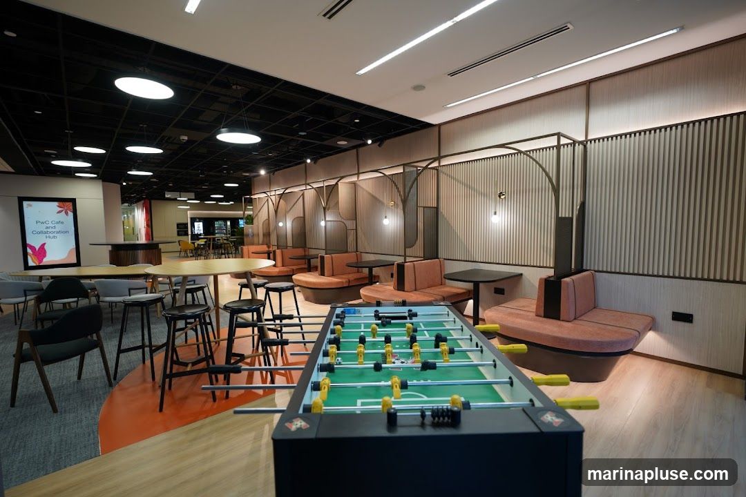 An interior view of a modern and spacious collaboration hub, likely a cafe or lounge area. The space features a variety of seating arrangements, including round tables with stools and comfortable booth-style seating with orange upholstery. A foosball table is prominently placed in the foreground, suggesting a recreational aspect to the space. The walls are decorated with vertical wooden slats and subtle lighting. In the background, a digital display shows "PwC Cafe and Collaboration Hub." The overall ambiance is contemporary and inviting, designed for both work and leisure.