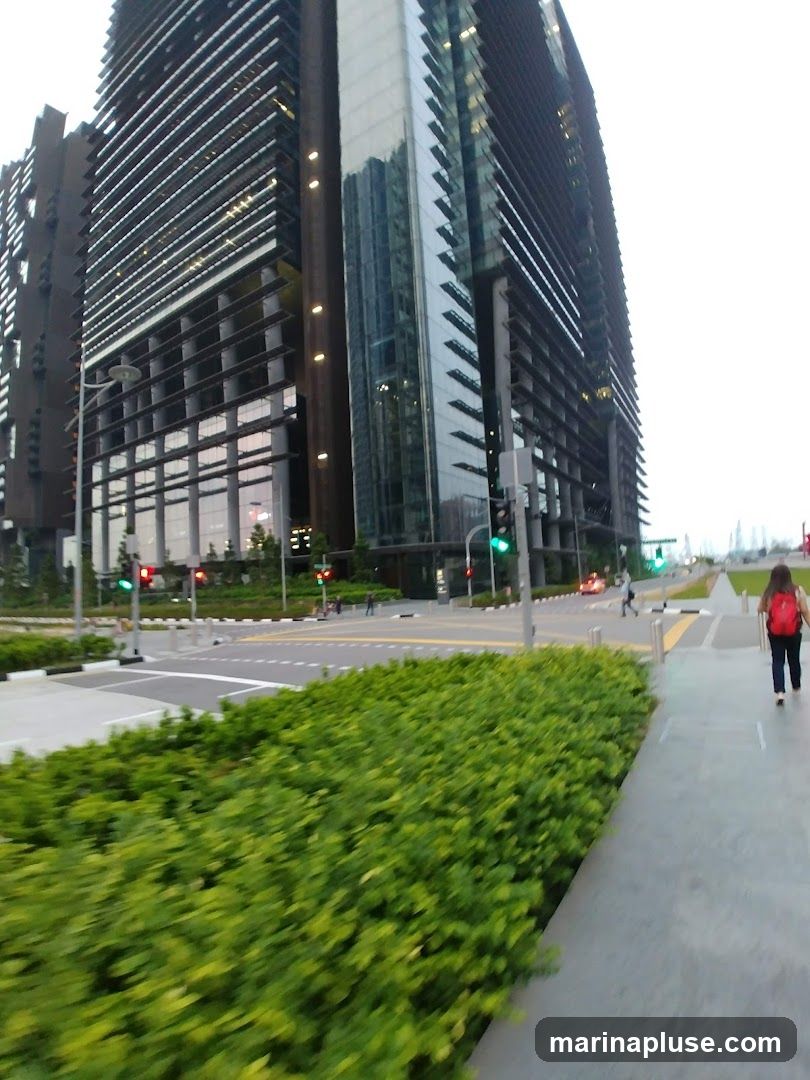 A wide shot of several modern, tall office buildings with glass and metal facades. In the foreground, there is a lush green hedge and a paved walkway. A person with a red backpack is walking away on the walkway. Traffic lights and a street are visible in the mid-ground. The sky is overcast.