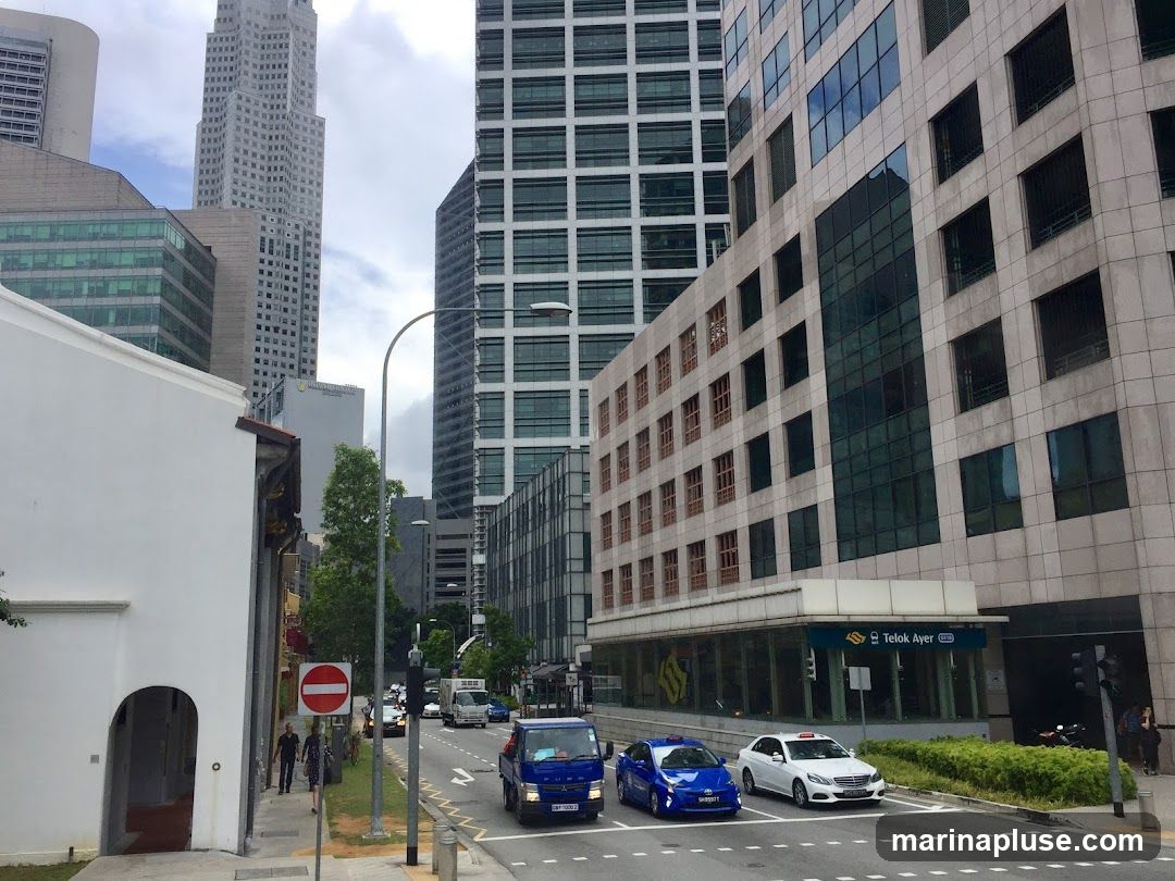 A street scene in a city with modern skyscrapers and a lower-rise building. Cars and a truck are driving on the road. A sign for 'Telok Ayer' is visible on one of the buildings. The sky is overcast.