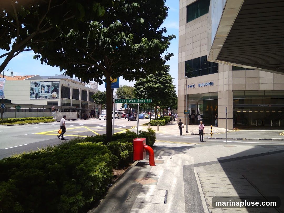 A view of the PWC Building entrance on a sunny day. The building has a modern facade with large windows. The words "PWC BUILDING" are visible in blue lettering above the entrance. People are seen entering and exiting the building. A street sign for "Cheang Wan Seng Pl" is visible in the foreground.
