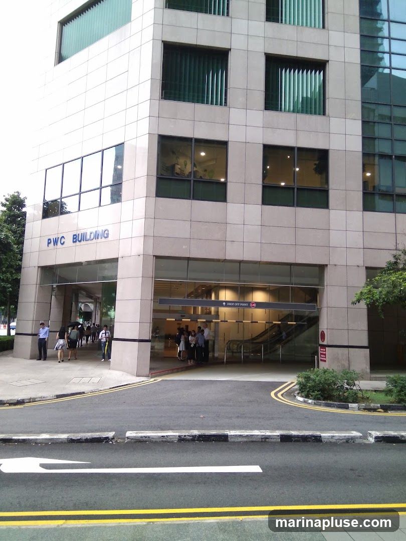 The exterior of the PWC Building shows a modern facade with large windows and a covered entrance. The building name, "PWC BUILDING," is visible in blue lettering on the side of the building. People are seen entering and exiting the building through the main entrance, which leads to escalators and an interior lobby. A sign indicates "DROP-OFF POINT." The street in front of the building has yellow and white lane markings.