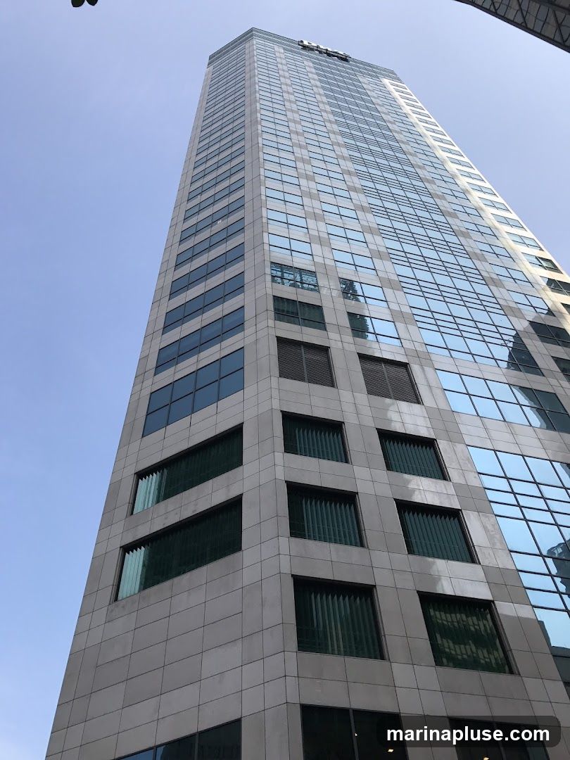 A low-angle shot of a modern skyscraper with a glass facade and stone accents. The building has many windows, some of which are covered by blinds. The sky is clear and blue.