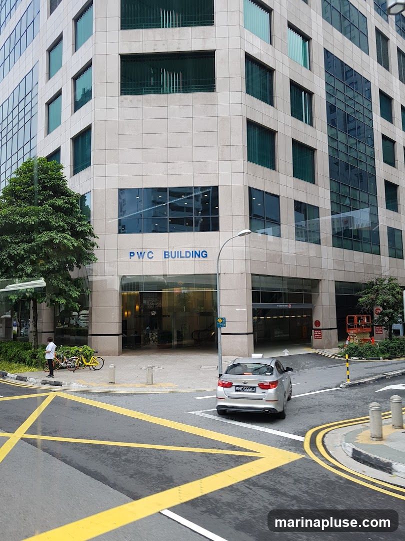 A view of the PWC Building's entrance on a street corner. The building has a modern facade with beige stone tiles and large glass windows. The words "PWC BUILDING" are visible in blue lettering on the building's exterior. A silver car is turning at the intersection in front of the building. Yellow road markings are prominent. A person stands near two yellow bicycles on the sidewalk.