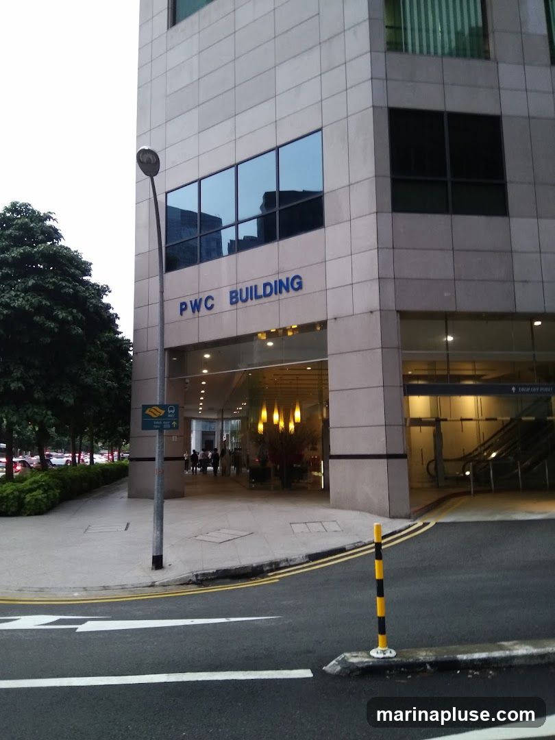 The exterior of the PWC Building is shown, with the building name prominently displayed in blue letters above the entrance. The entrance area features large glass windows and doors, with interior lighting visible. To the left, a street lamp and a bus stop sign are present. To the right, an escalator leading to a drop-off point is visible. The building has a modern facade with large windows reflecting the sky and surrounding structures.