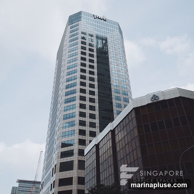 A low-angle shot of two modern office buildings against a cloudy sky. The taller building on the left has a glass facade with a visible logo at the top. The building on the right has a darker glass facade and features a logo and text that reads "SINGAPORE OFFICE SPACES" on its side. A construction crane is visible in the background.