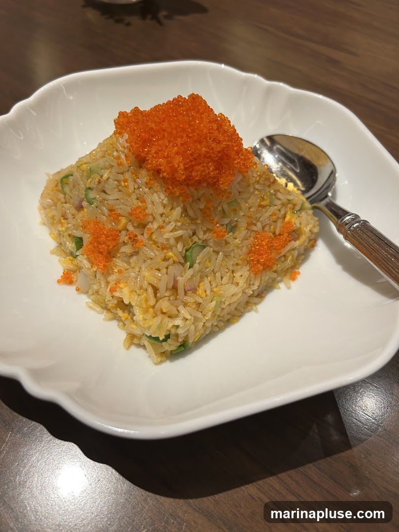 A serving of fried rice topped with bright orange tobiko (flying fish roe) is presented in a white, scalloped-edge bowl. A spoon rests on the right side of the bowl. The fried rice appears to contain small pieces of vegetables and egg. The dish is placed on a dark wooden table.