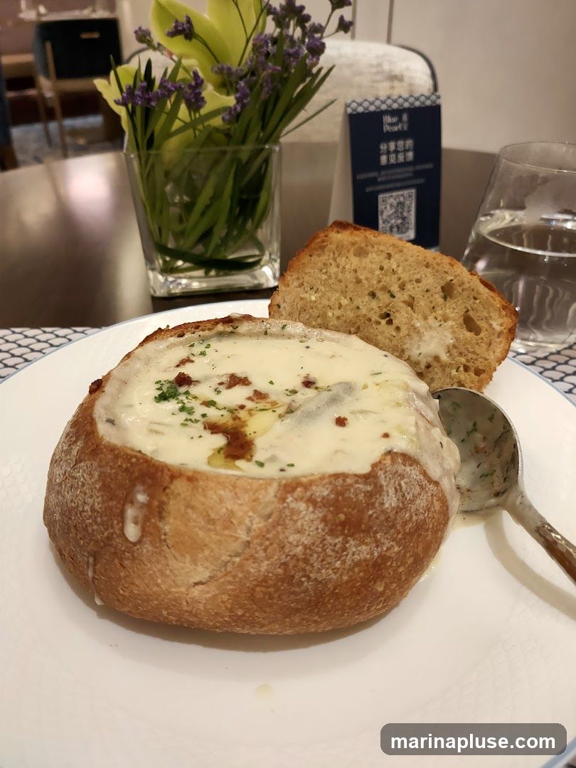 A close-up shot of a bread bowl filled with creamy soup, topped with herbs and a drizzle of oil. A slice of bread sits on the side of the bowl, and a spoon rests on the plate. A small vase of flowers and a menu card are visible in the background.