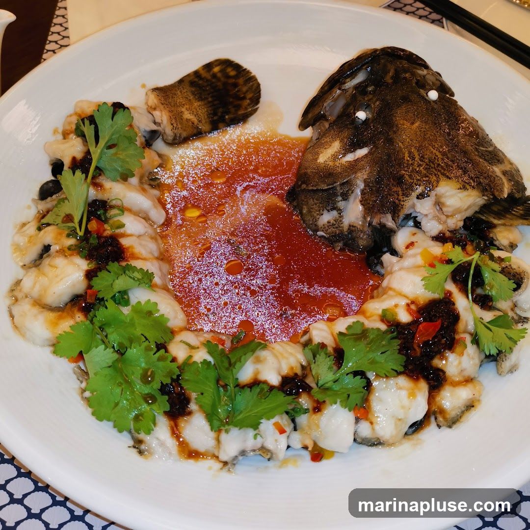 A white plate holds a whole steamed fish, with the head and tail sections prominently displayed. The fish is arranged in slices around the head, covered in a savory sauce with chili, black beans, and garnished with fresh cilantro. The dish is presented on a table with a patterned placemat and chopsticks in the background.