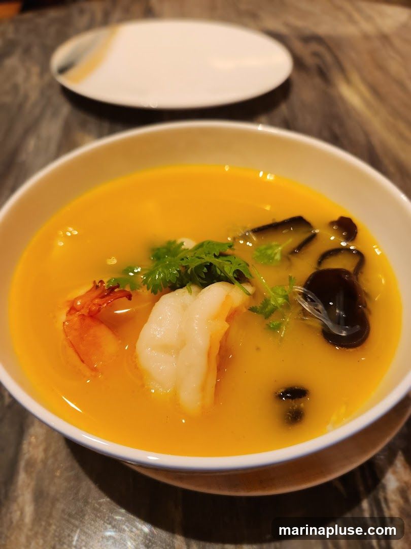A close-up shot of a bowl of soup with a bright orange broth. Visible ingredients include a large shrimp, pieces of dark mushroom, and fresh cilantro. A white plate with a decorative rim is blurred in the background. The soup is served in a white bowl on a textured surface.
