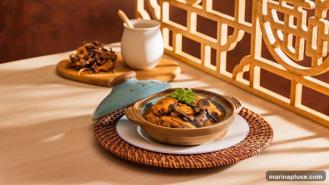 A close-up shot of a steaming clay pot filled with a savory dish, likely chicken and mushrooms, garnished with fresh cilantro. The pot is placed on a woven placemat on a light-colored wooden table. In the background, a small white pitcher with a wooden spoon sits next to a pile of dried mushrooms on a wooden board. A decorative wooden screen with an intricate lattice pattern is visible on the right side of the image, casting shadows on the table.