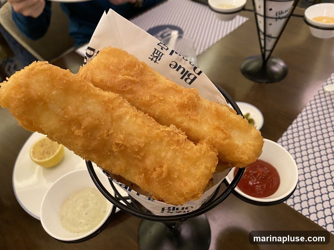 Two pieces of battered fish served with a lemon wedge, tartar sauce, and ketchup. The fish is presented in a black wire basket lined with paper that has a blue logo.