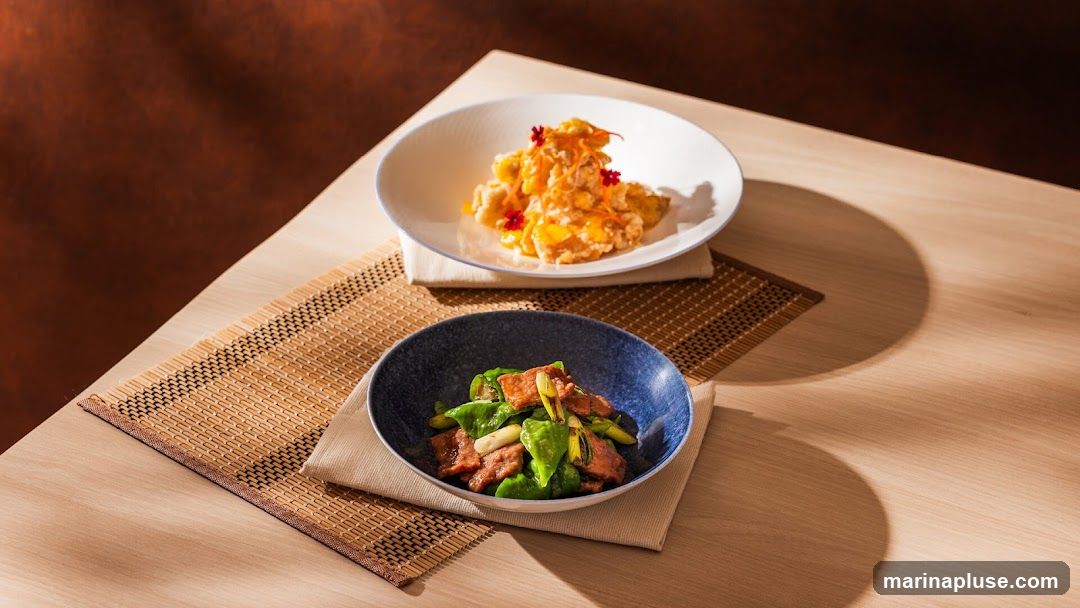 Two bowls of food are presented on a wooden table. The bowl in the foreground contains sliced meat with green peppers and scallions. The bowl in the background contains a dish with a yellow sauce, possibly fried chicken or fish, garnished with shredded carrots and red flowers.