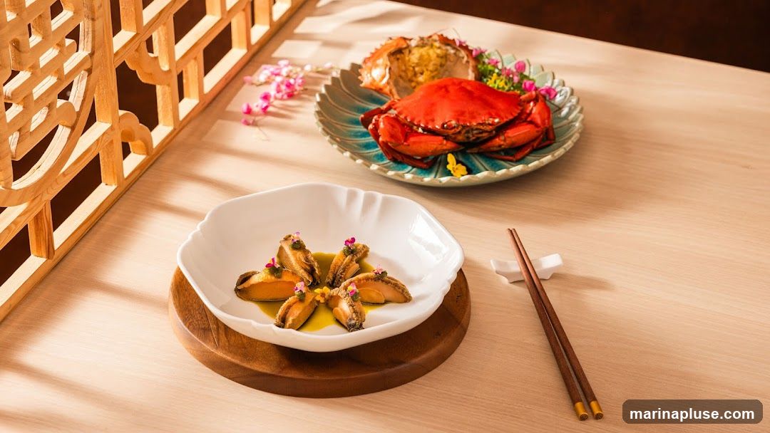 A white, scalloped-edge bowl holds several pieces of abalone, each topped with a dollop of green sauce and a small pink flower. The bowl sits on a round wooden coaster. In the background, a whole cooked crab is presented on a decorative blue and green plate, garnished with pink and yellow flowers. Chopsticks rest on a white chopstick holder to the right of the abalone dish. The setting appears to be a restaurant table with a light wood grain surface, and a decorative wooden screen is visible on the left side of the image.