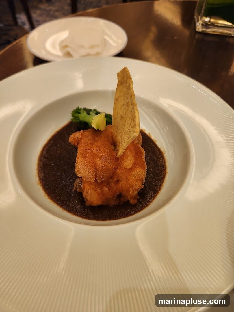 A close-up shot of a white plate with a serving of fried fish on top of a dark mole sauce. A piece of broccoli and a lime wedge are visible next to the fish, and a tortilla chip is placed on top of the fish. A small white dish with what appears to be rice or noodles is in the background. The table surface is a reflective, dark wood.