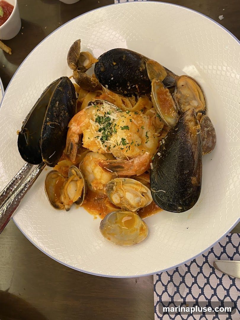 A white plate filled with pasta, mussels, clams, and shrimp in a tomato-based sauce, topped with grated cheese and parsley.