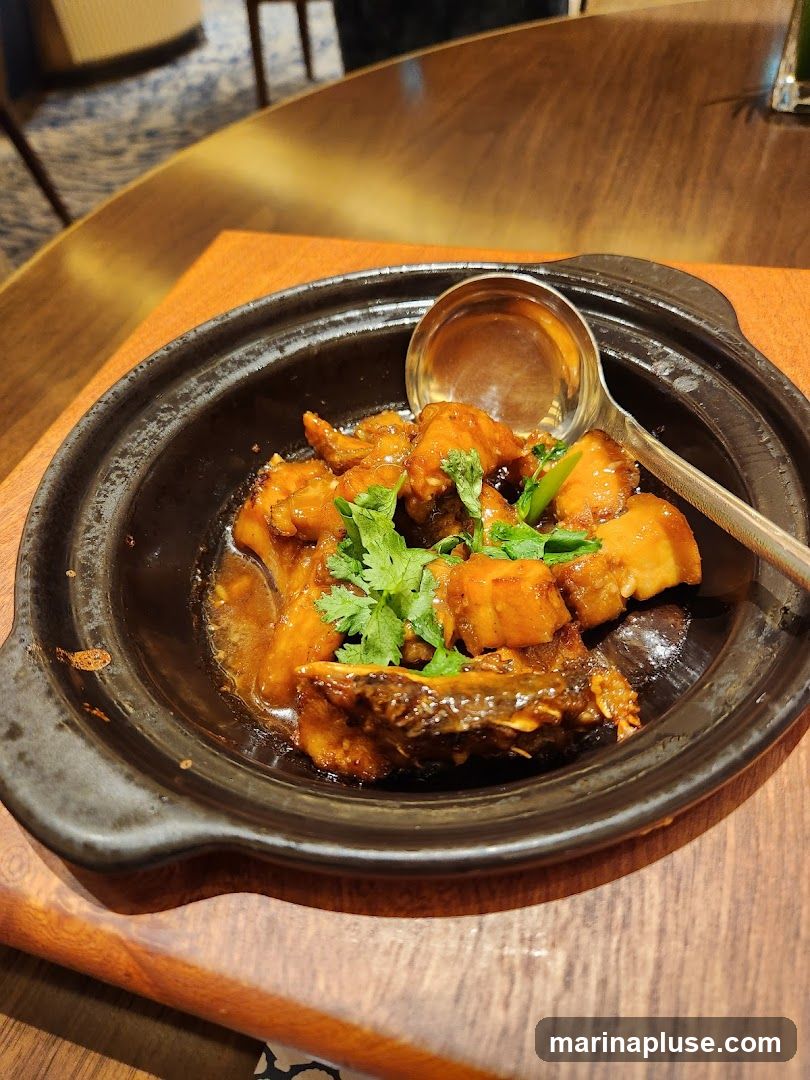 A close-up shot of a braised fish dish served in a dark ceramic pot. The fish pieces are coated in a glossy brown sauce and garnished with fresh cilantro. A ladle rests on the side of the pot. The dish is placed on a wooden surface, with a blurred background suggesting a restaurant setting.
