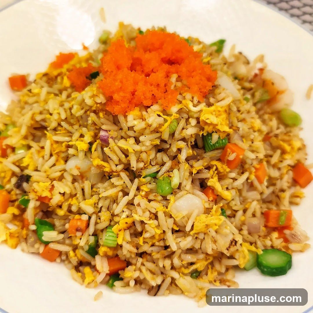 A close-up shot of a plate of fried rice topped with bright orange tobiko (flying fish roe). The fried rice contains visible pieces of scrambled egg, diced carrots, peas, chopped green onions, and what appear to be small pieces of seafood like shrimp or scallops. The dish is served on a white plate.