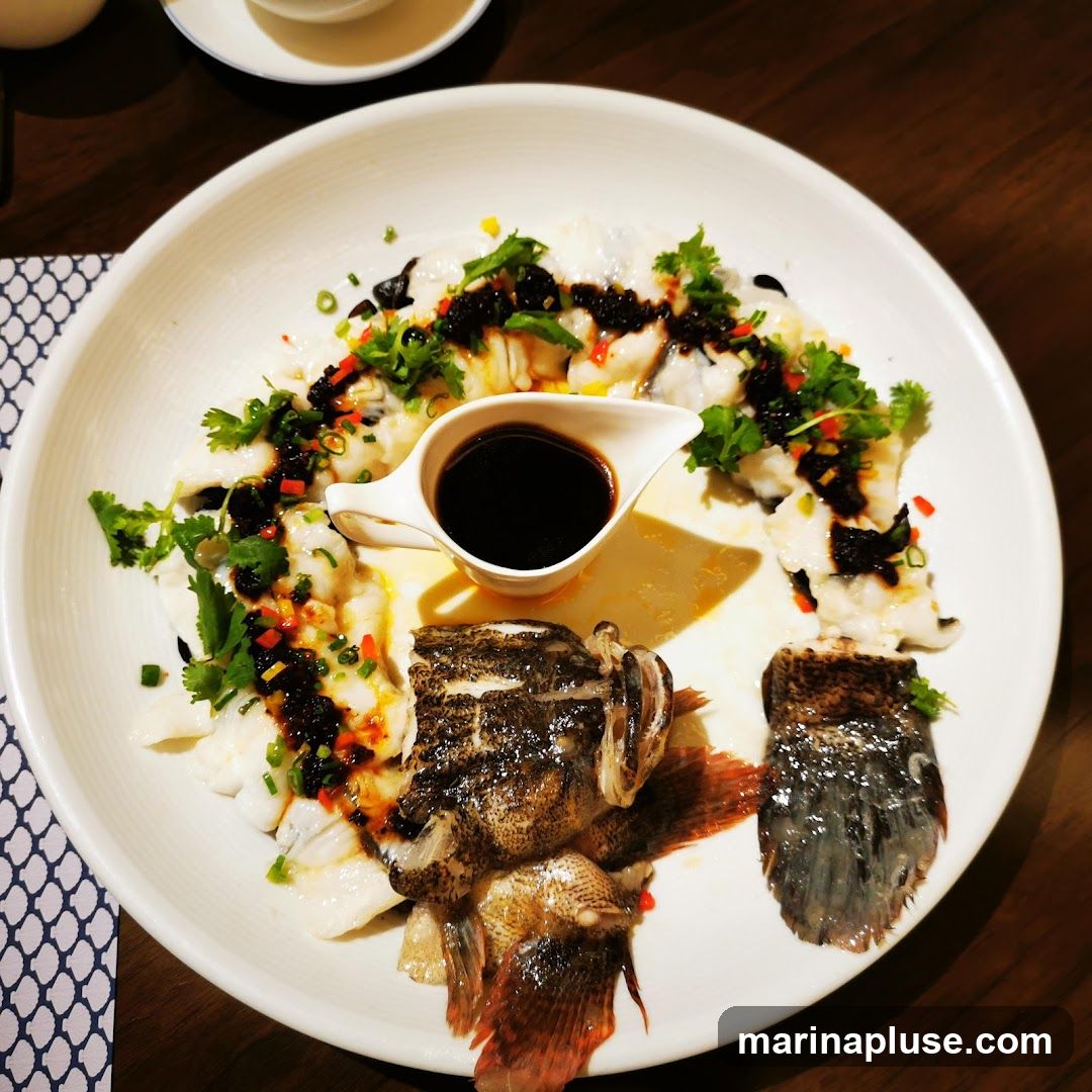 A white plate holds a steamed fish dish. The fish is arranged in pieces, topped with a dark sauce, chopped scallions, cilantro, and chili flakes. A small white dish of dark sauce is in the center. The head and tail of the fish are also on the plate.