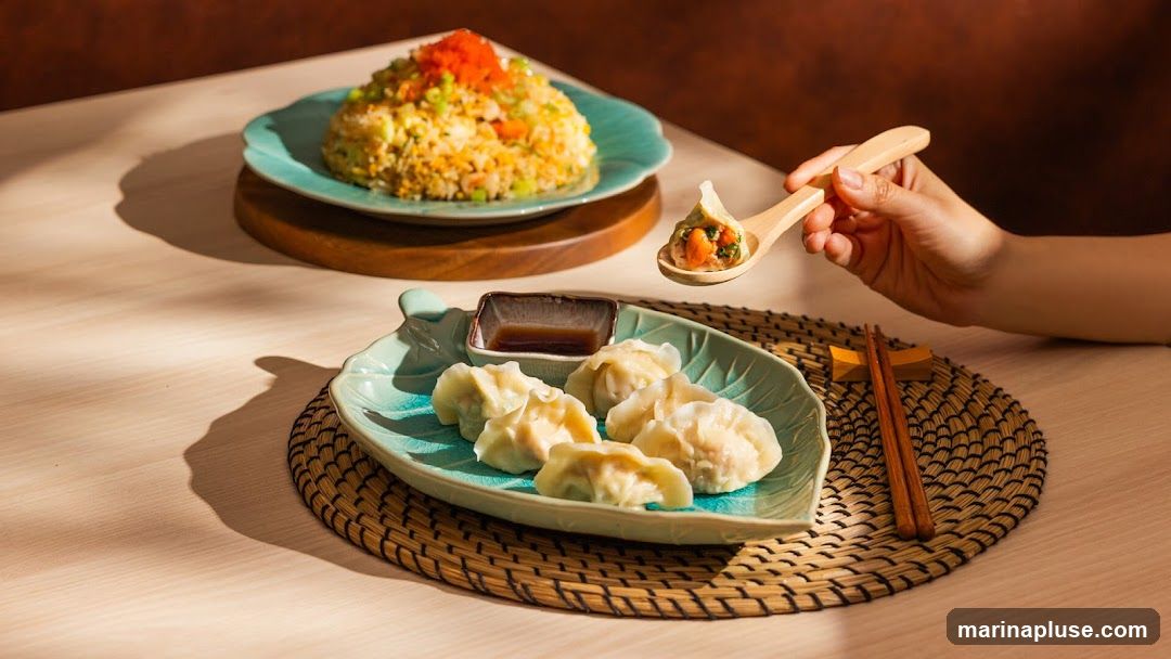 A close-up shot of a meal featuring dumplings and fried rice. A hand is holding a wooden spoon with a single dumpling filled with ingredients. The dumplings are served on a light blue, leaf-shaped plate with a small dish of dipping sauce. In the background, a mound of fried rice is presented on another blue plate. The food is arranged on a wooden table with a woven placemat.