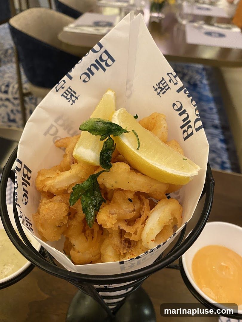 A cone of fried calamari is served with lemon wedges and garnished with fresh herbs. Two small bowls of dipping sauce are placed on either side of the cone. The paper lining the cone has the text "Blue Door" printed on it. The background shows a blurred restaurant interior with tables and chairs.