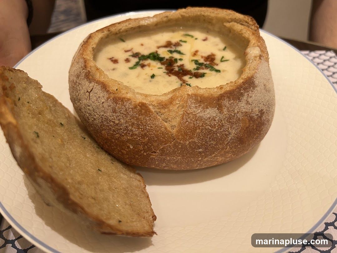 A close-up shot of a creamy soup served in a hollowed-out bread bowl. The soup is garnished with chopped green herbs and small pieces of bacon. A slice of toasted bread, seasoned with herbs, is placed on the side of the bowl on a white plate with a subtle textured pattern.