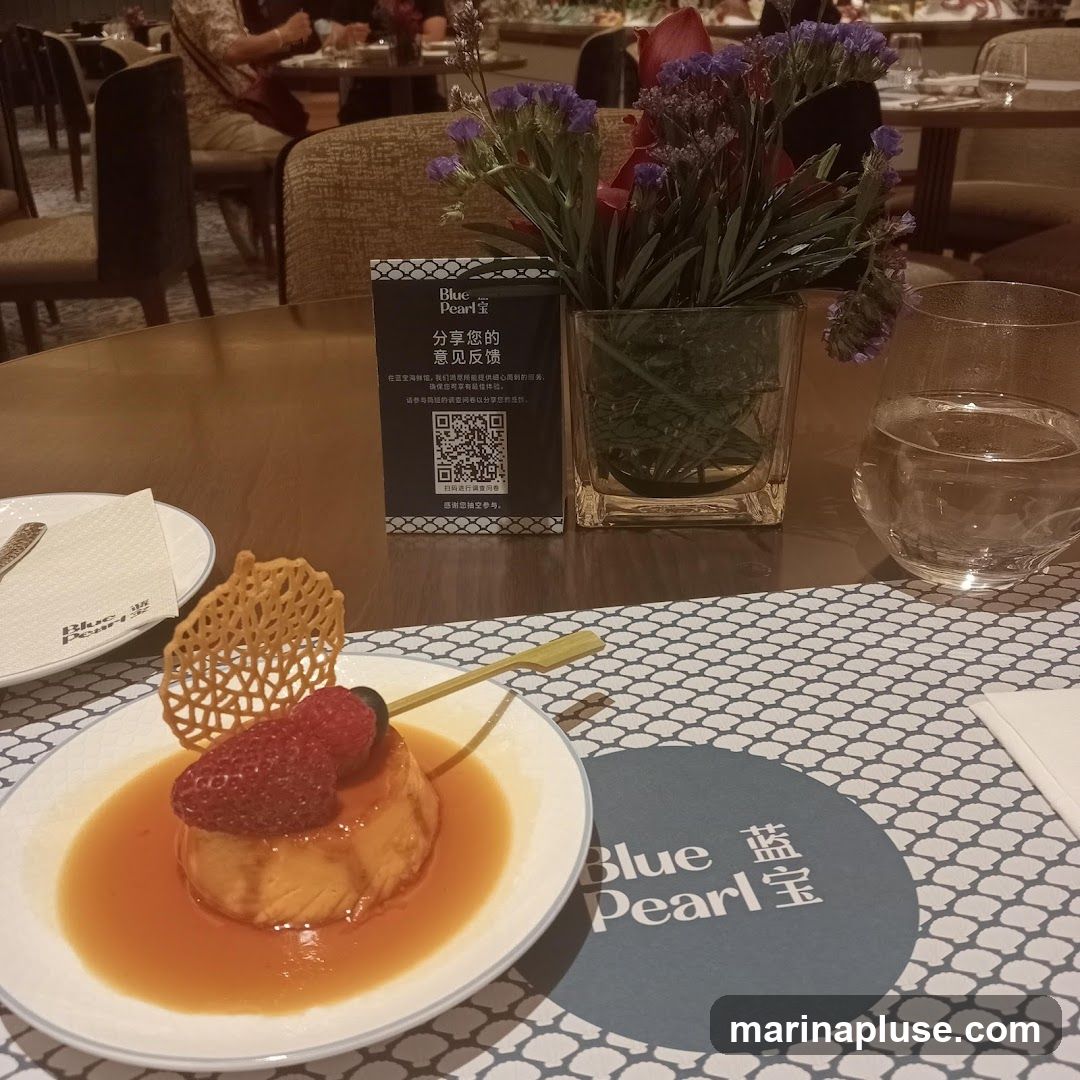 A close-up view of a dessert, possibly a crème caramel, topped with strawberries and a blueberry, served on a white plate with a decorative crisp. The dessert is placed on a table with a patterned placemat. A small sign with Chinese text and a QR code, along with a vase of flowers and a glass of water, are also visible. The branding "Blue Pearl" is present on the placemat and the sign.