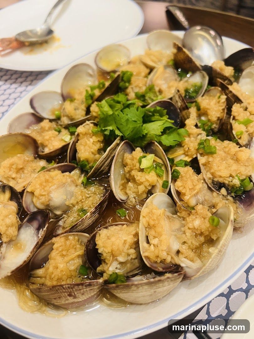 A close-up shot of a platter of steamed clams, generously topped with minced garlic and chopped scallions. The clams are served in a light broth with thin noodles. A sprig of cilantro garnishes the dish. In the background, a white plate with a spoon and a shrimp tail is visible.