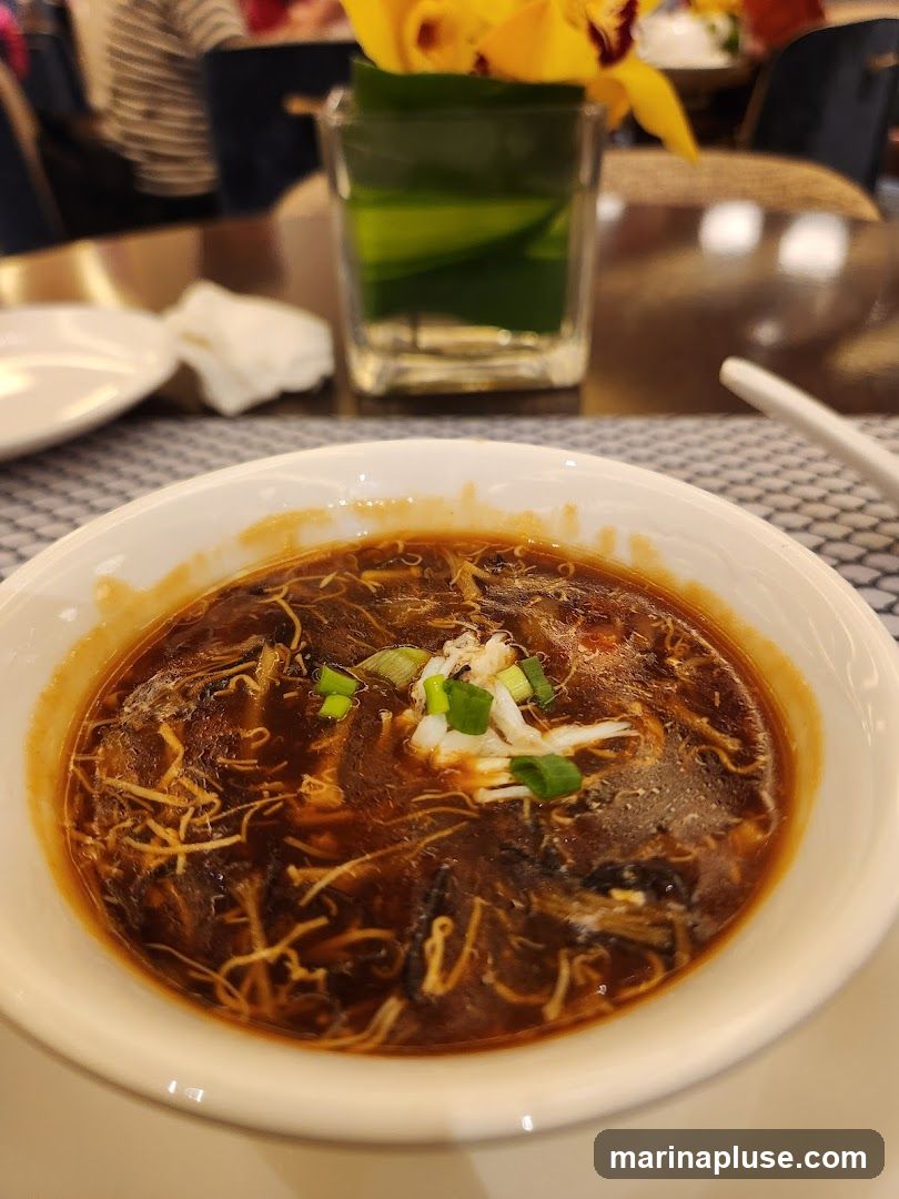 A close-up shot of a bowl of hot and sour soup, garnished with chopped green onions and shredded white meat. The soup is a deep brown color with visible strands of ingredients. In the background, a glass vase with yellow orchids and green leaves sits on a table, along with a white napkin and a plate. The setting appears to be a restaurant.