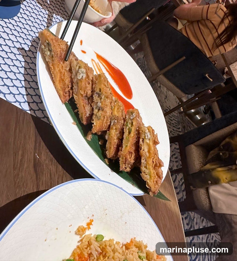A plate of fried appetizers, possibly stuffed with seafood and vegetables, is served with a drizzle of red sauce. Another plate in the foreground contains fried rice with peas and roe. The setting appears to be a restaurant with people dining in the background.