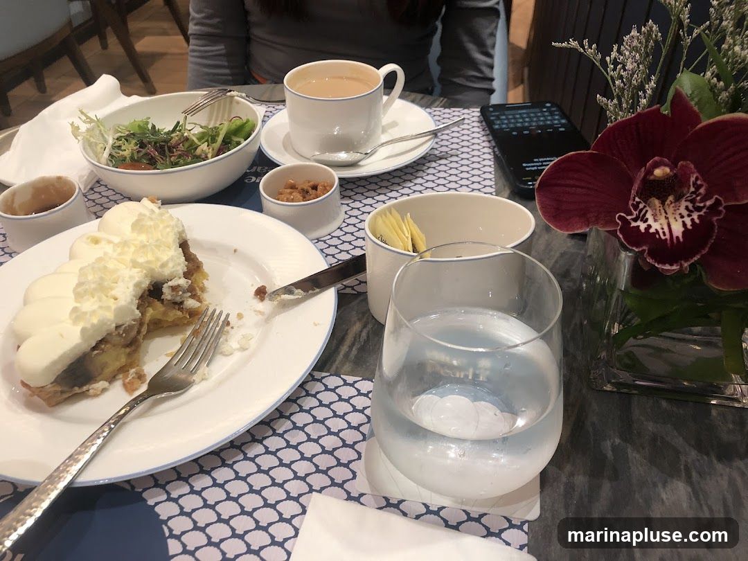 A slice of tart with whipped cream, a salad, a cup of tea, and a glass of water are arranged on a table. A dark red orchid is in a vase on the right. A smartphone is also visible on the table.