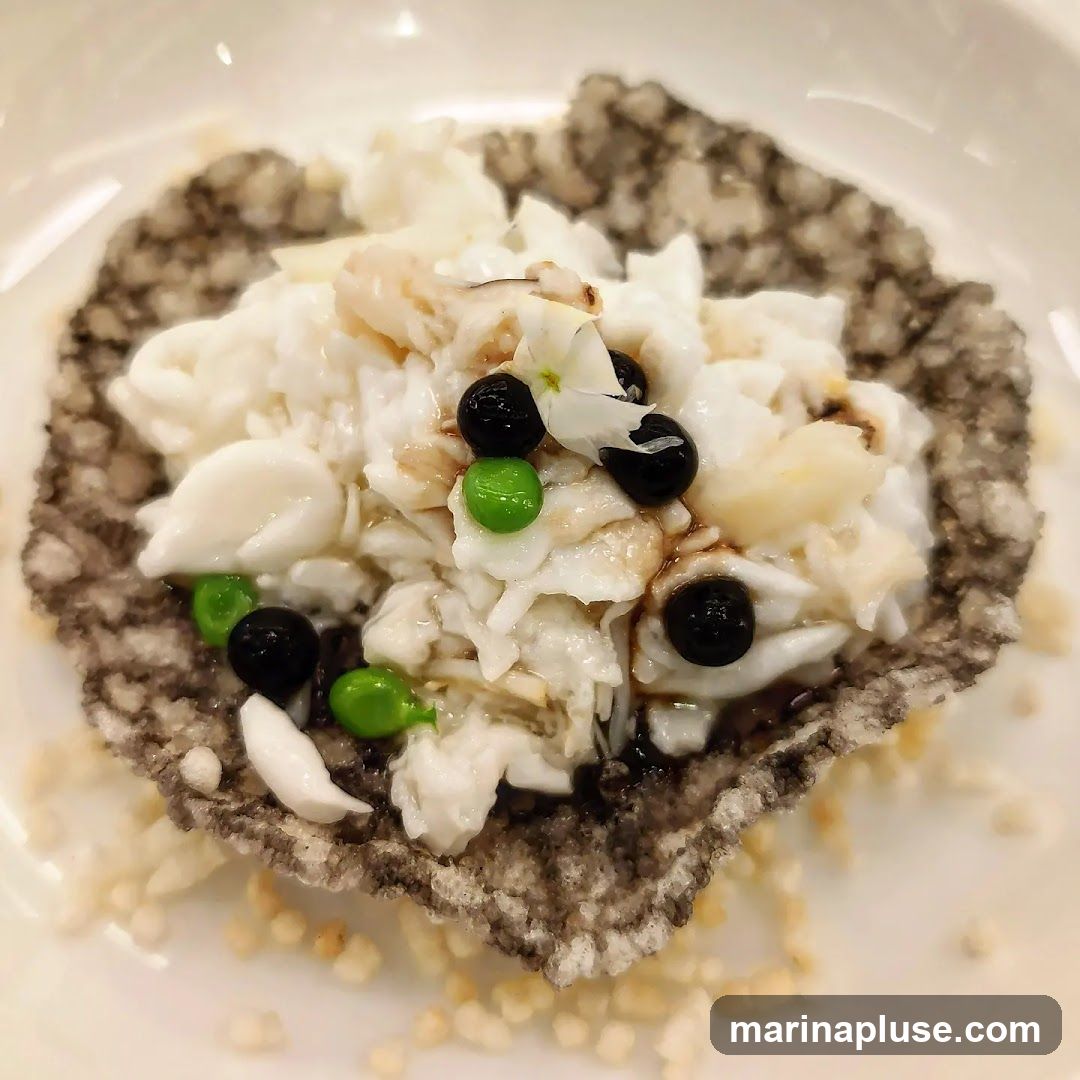 A close-up shot of a dish featuring shredded crab meat, bright green peas, and black beans, served on a crispy, dark-colored cracker. The dish is garnished with a small white flower and drizzled with a dark sauce. The cracker is placed on a bed of puffed rice or similar crispy grains.