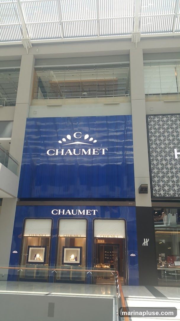 The exterior of a Chaumet jewelry store is shown, featuring a large blue facade with the brand name and logo prominently displayed. Below this, the storefront has three large display windows showcasing jewelry. The entrance is visible in the center.