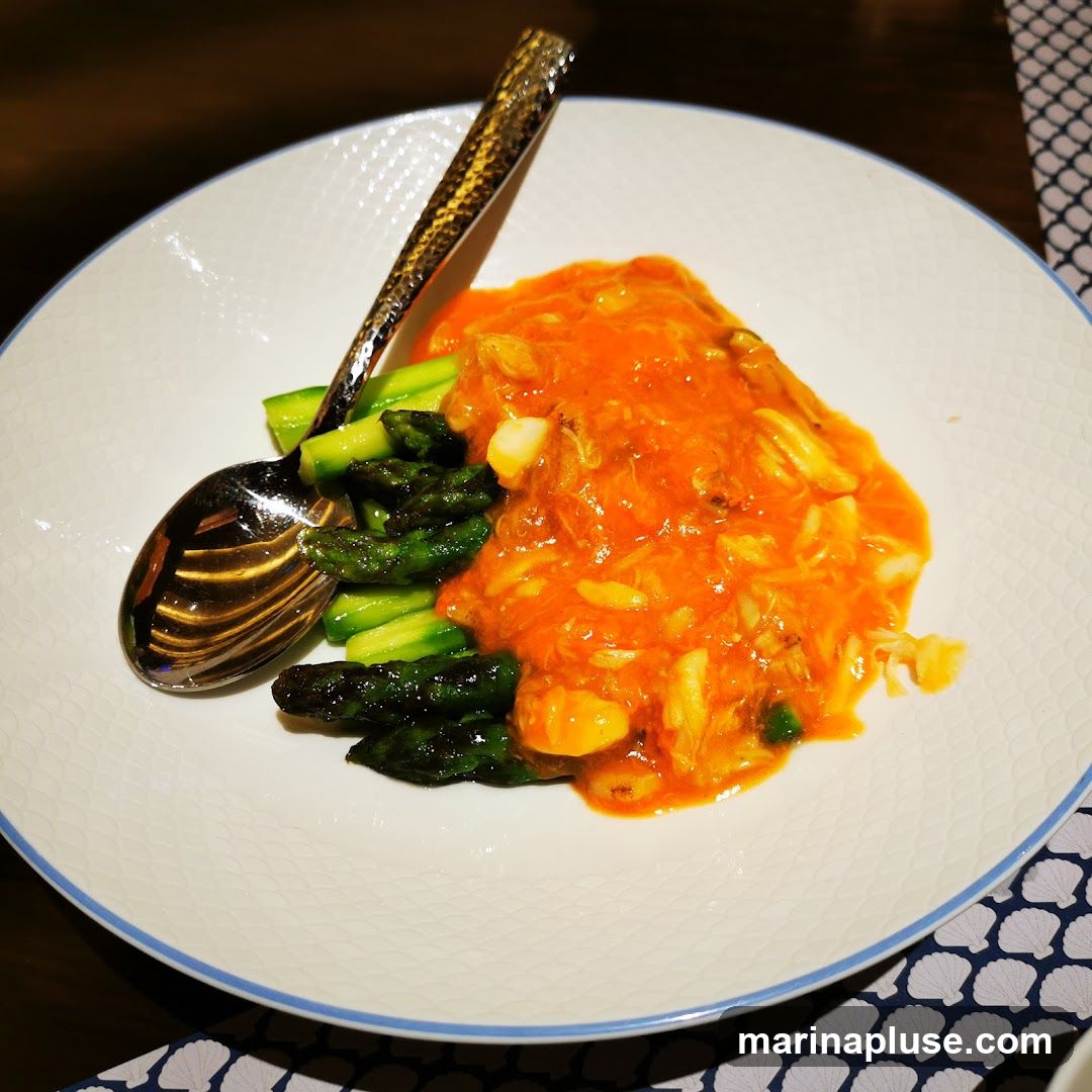 A white plate with a blue rim holds a dish of asparagus spears topped with a vibrant orange sauce containing shredded crab meat. Two spoons are placed on the plate, one resting on the asparagus and the other angled towards the top of the dish. The plate is set on a dark surface with a patterned placemat.