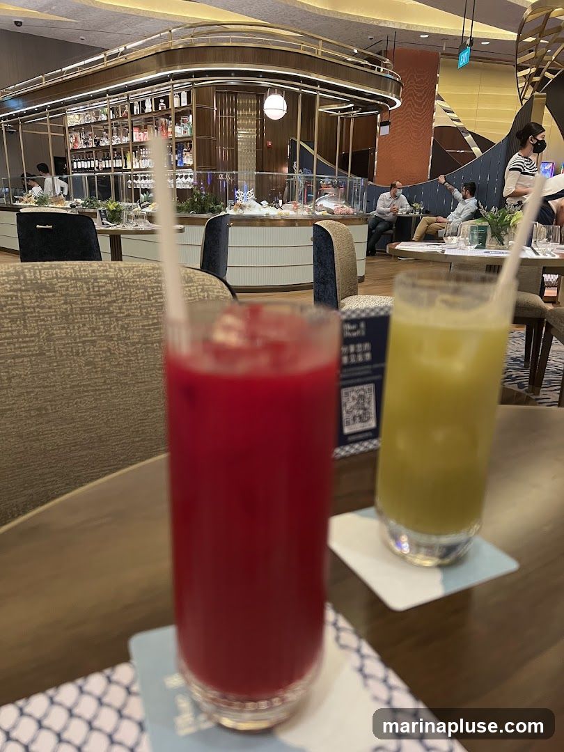 Two tall glasses of colorful drinks, one red and one green, sit on a table in front of a bar area. The bar is well-stocked with bottles and glassware, and there are people seated at tables in the background. The overall ambiance appears to be upscale and modern.