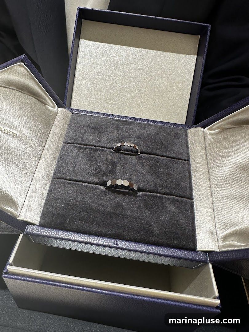 An open jewelry box with two rings inside. The box is dark blue with a textured exterior and a satin lining. The interior has a dark gray velvet lining with indentations for the rings. One ring appears to be a band with small diamonds, and the other is a geometric band.