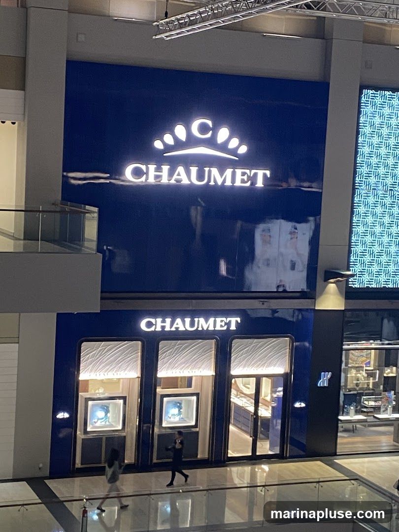 The exterior of a Chaumet jewelry store. The store has a dark blue facade with the Chaumet logo and name displayed prominently in white, illuminated lettering. The storefront features large display windows showcasing jewelry. Two individuals are walking past the entrance.