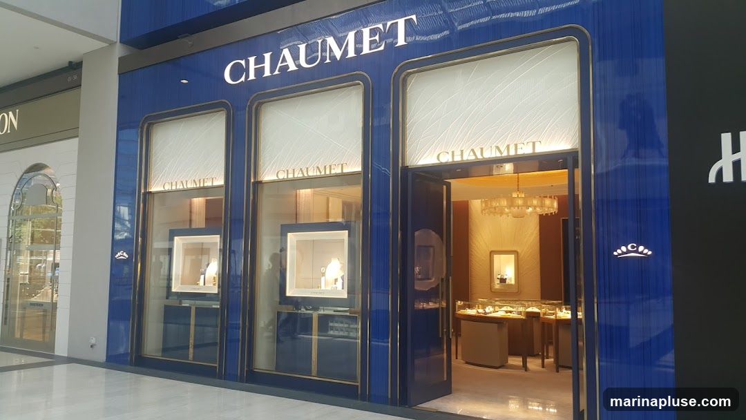 The exterior of a luxury jewelry store, Chaumet, with a blue facade and gold trim. The store features large display windows showcasing jewelry. The entrance is open, revealing the interior with display cases and a chandelier. The brand name "CHAUMET" is prominently displayed above the windows.