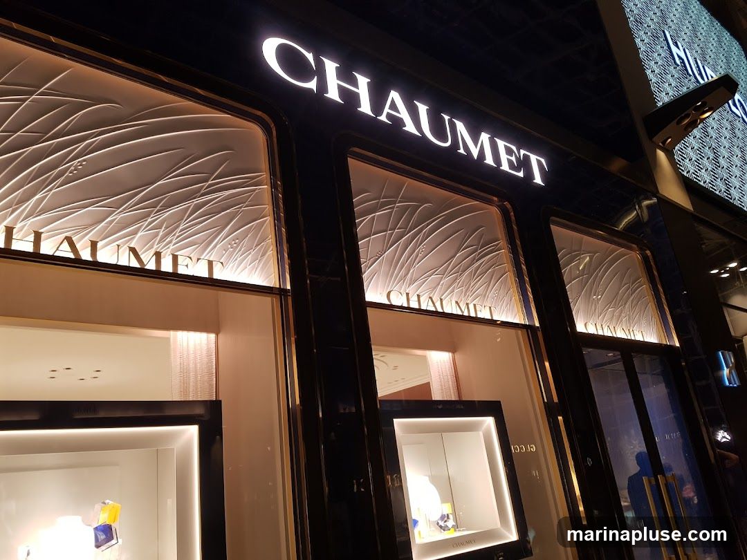 A nighttime exterior view of a luxury jewelry store. The prominent "CHAUMET" logo is illuminated in large white letters above the storefront windows. The windows display intricate, wave-like patterns illuminated from behind, with some display cases visible inside. The facade is dark and sleek, with gold trim around the windows.