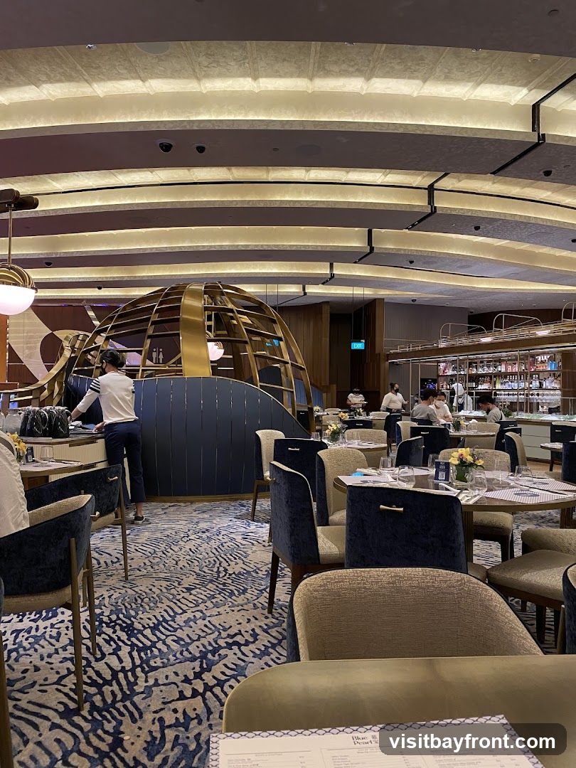 The image shows the interior of a restaurant with a modern and elegant design. The dining area features round tables set with white tablecloths and chairs upholstered in dark blue and beige fabric. A distinctive golden, spherical structure is visible in the background, along with a bar area stocked with bottles. The ceiling has a unique curved design with integrated lighting. Staff members are seen attending to tables and working behind the bar. The floor is covered with a patterned carpet in shades of blue and beige. A menu is partially visible in the foreground.