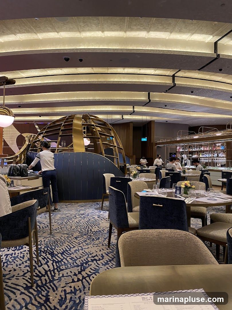 The image shows the interior of a restaurant with a modern and elegant design. The dining area features round tables set with white tablecloths and chairs upholstered in dark blue and beige fabric. A distinctive golden, spherical structure is visible in the background, along with a bar area stocked with bottles. The ceiling has a unique curved design with integrated lighting. Staff members are seen attending to tables and working behind the bar. The floor is covered with a patterned carpet in shades of blue and beige. A menu is partially visible in the foreground.