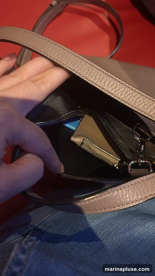 A hand is reaching into a light brown pebbled leather purse. Inside the purse, a small zippered pouch, a blue object, and a black item are visible. The purse has a strap and is resting on a red surface, with denim fabric visible at the bottom.