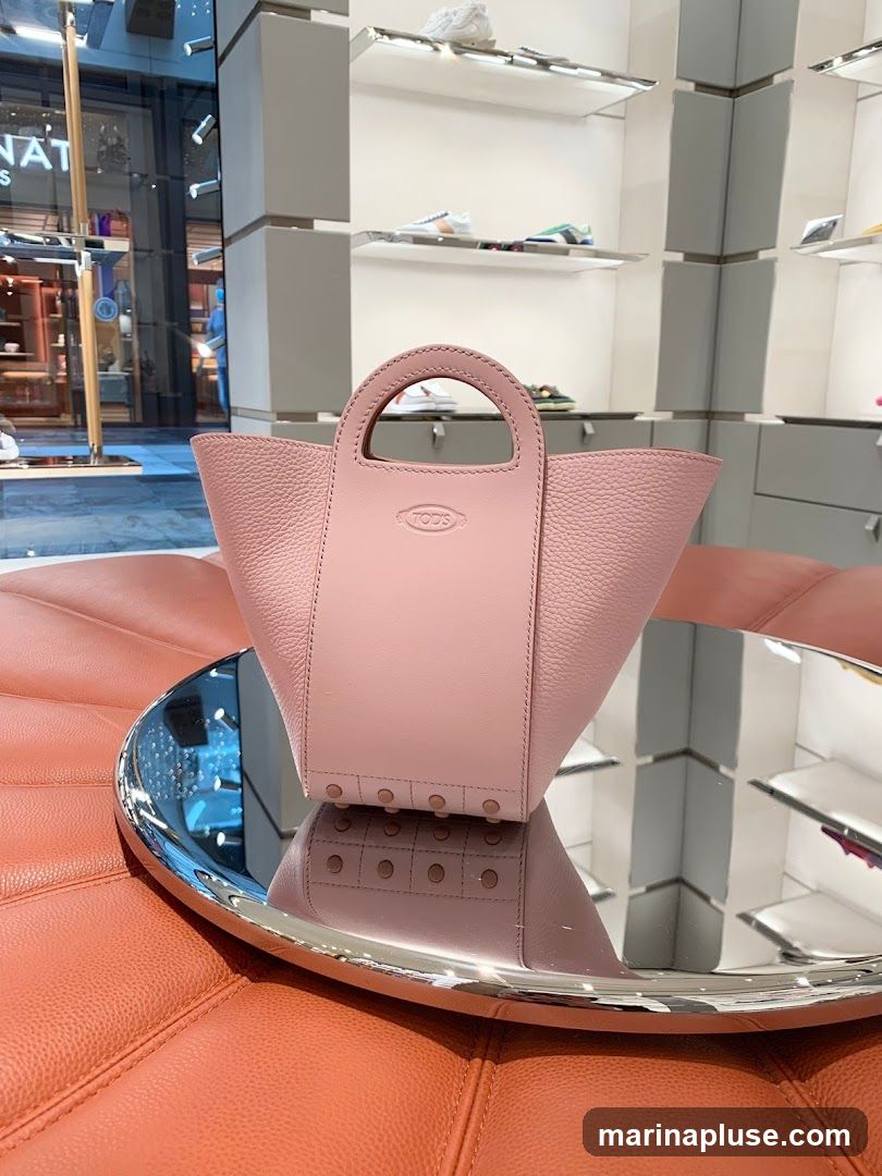A pink Tod's handbag with a pebbled leather finish is displayed on a mirrored tray. The handbag has a structured design with a central panel featuring the embossed Tod's logo. The tray reflects the handbag and the interior of a store, which has shelves displaying shoes. The background shows a store interior with display shelves and lighting.