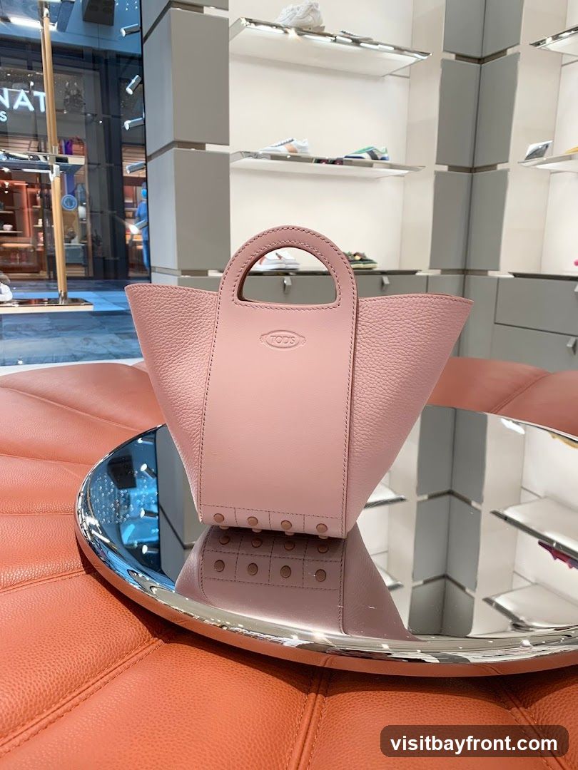 A pink Tod's handbag with a pebbled leather finish is displayed on a mirrored tray. The handbag has a structured design with a central panel featuring the embossed Tod's logo. The tray reflects the handbag and the interior of a store, which has shelves displaying shoes. The background shows a store interior with display shelves and lighting.