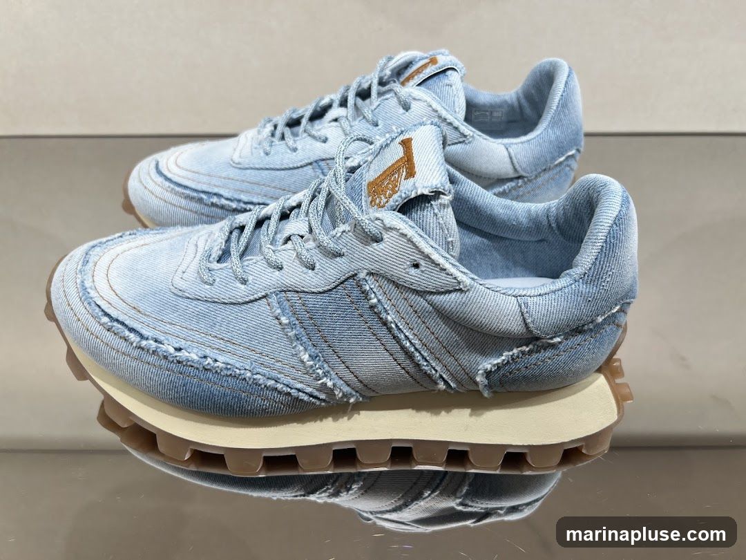 A pair of light blue denim sneakers with frayed edges and tan laces are displayed on a reflective surface. The sneakers have a tan logo embroidered on the tongue.